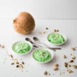 Three bowls filled with green pistachio pudding, topped with crushed pistachios, surrounded by three spoons, a coconut and more crushed pistachios.
