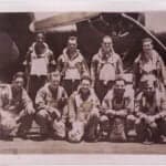 Sepia-toned group portrait of ten men in flight suits and harnesses, with an airplane propeller and engine in the background. Four men stand in the back row, and six men crouch in the front row. Handwritten text is visible vertically on the right edge of the photograph.