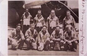 Sepia-toned group portrait of ten men in flight suits and harnesses, with an airplane propeller and engine in the background. Four men stand in the back row, and six men crouch in the front row. Handwritten text is visible vertically on the right edge of the photograph.