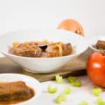 Close up - Two white bowls filled with Swiss Steak and gravy, surrounded by tomatoes, celery and an onion.