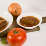 Two white bowls filled with Swiss Steak and gravy, surrounded by tomatoes, celery and an onion.