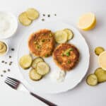 Top view- Two tuna patties on a white plate with pickle chips and lemon wedges.