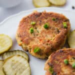 Close up-two tuna patties on a white plate with pickle chips.