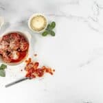 Top view - White bowl filled with Parmesan Meatballs, topped with marinara sauce. A fork with a meatball on it and a bowl of parmesan cheese to the side.