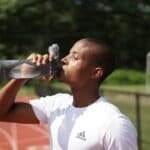 Dark grey BINO brand reusable water bottle with clear sections, held by a person drinking. Person wears a white t-shirt with a dark grey adidas logo.
