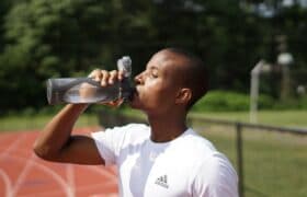 Dark grey BINO brand reusable water bottle with clear sections, held by a person drinking. Person wears a white t-shirt with a dark grey adidas logo.