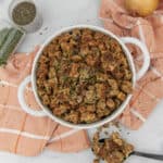 A white ceramic casserole dish holds golden-brown stuffing garnished with green herbs. A black serving spoon with a scoop of stuffing rests beside the dish. Two glass spice jars with dried herbs and two whole yellow onions are on a white marble countertop with an orange and white plaid towel.