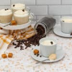 Six servings of coffee and cream dessert or drink are displayed. Four are in clear glass mugs on a white tiered stand, two in white ceramic mugs with saucers. Each serving is topped with white cream and caramel drizzle, garnished with coffee beans. An overturned clear glass jar has spilled dark roasted coffee beans onto a light mosaic counter. Loose coffee beans and orange caramel squares are scattered around. A white and orange striped kitchen towel lies on the counter. The background is a white subway tile wall.
