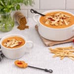 A large white serving bowl and a smaller white mug of orange-red soup are garnished with rectangular crackers, diced red tomatoes, and green herbs. Fresh cilantro and dill are in a glass jar, brown ground spice is spilled from a container, a small bowl holds diced tomatoes, and a vine of fresh tomatoes are present. A pile of crackers and a black and white patterned napkin complete the scene on a white cracked-texture surface.