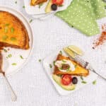 Overhead view of a round golden brown layered savory pie on a white plate, with a slice removed by a silver spatula. A serving of the pie on a white square plate features a dollop of white cream, a black olive, sliced avocado, a tomato wedge, and green onion garnish. Another partial slice is visible on a second white plate. A green polka dot napkin, a bowl of red spices, and scattered lime wedges are also arranged on a white crackled surface.