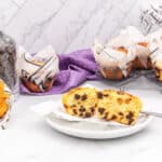 Chocolate chip muffins in newspaper print liners are displayed on a white surface with a purple polka dot cloth. One muffin is cut in half on a small white plate, revealing its chocolate chip interior. A clear glass jar filled with dark chocolate chips and a white scoop is visible on the left.