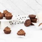 Chocolate brownie bites or mini muffins, some plain dark brown, others topped with light brown frosting and a single coffee bean, arranged on a white cake stand, a white marble-patterned surface, and in white paper liners, with a white and black polka dot fabric visible.