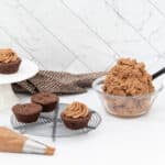 Chocolate cupcakes, one frosted on a white cake stand, three plain on a cooling rack. A glass bowl of light brown frosting with a black spatula, and a piping bag with frosting and a metal tip are on a white marble surface. A brown polka dot cloth is draped behind the cake stand.