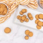 Overhead view of a white rectangular plate displaying many light brown peanut butter cups, some in dark fluted liners and some without. A white bowl of creamy peanut butter, a clear jar of whole peanuts, and a striped yellow and white napkin are also on a white marble surface.