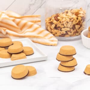Tan peanut butter cups with dark brown bases are arranged on a white marble surface. Some are on a white rectangular plate, some are stacked, and one is in a fluted paper wrapper. A clear glass jar of whole peanuts and a white bowl of peanut butter are in the background, next to a yellow and white striped towel.
