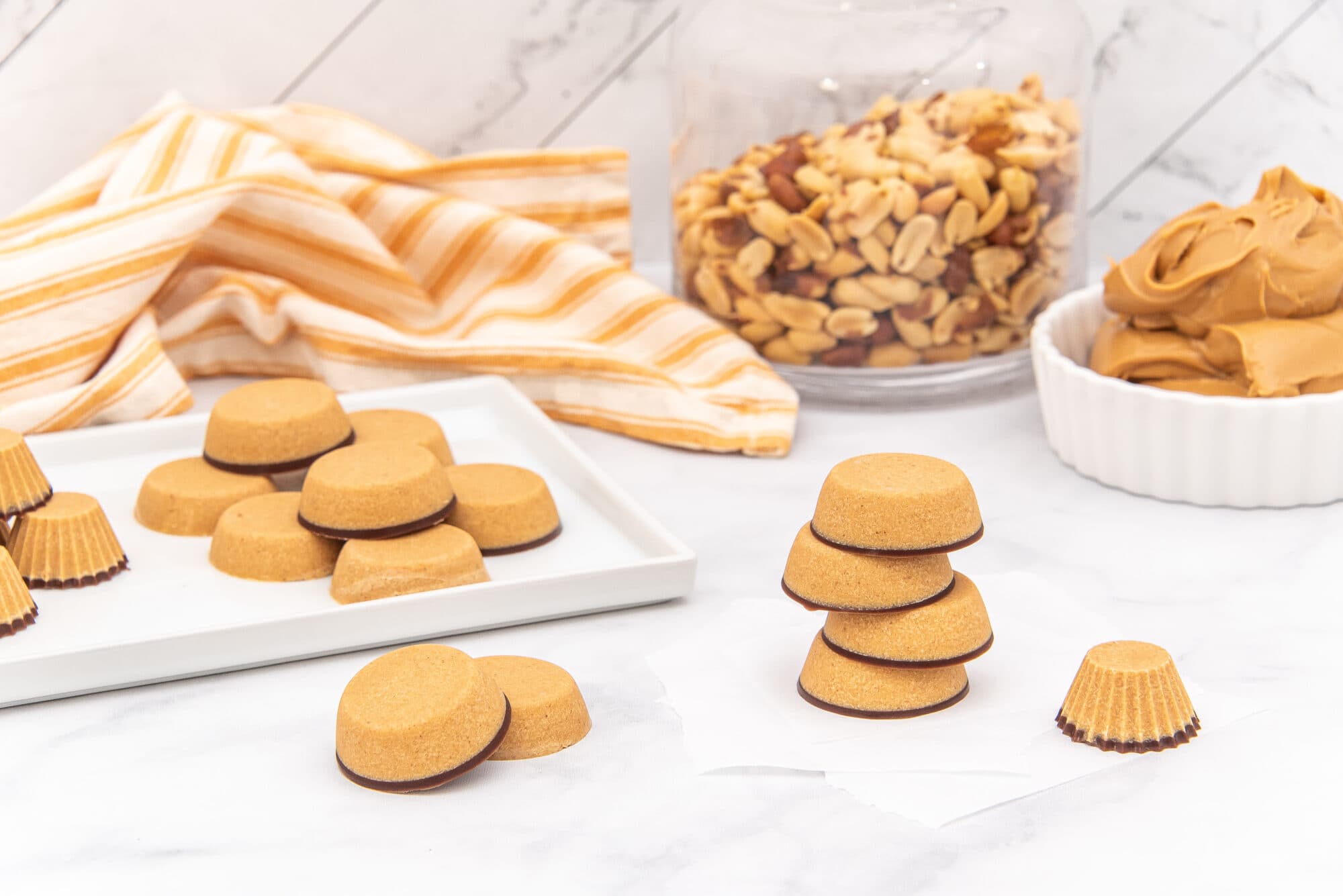 Tan peanut butter cups with dark brown bases are arranged on a white marble surface. Some are on a white rectangular plate, some are stacked, and one is in a fluted paper wrapper. A clear glass jar of whole peanuts and a white bowl of peanut butter are in the background, next to a yellow and white striped towel.
