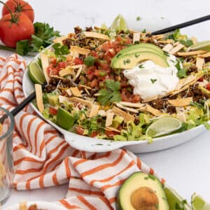 Large white bowl of taco salad containing ground beef, shredded lettuce, diced tomatoes, shredded cheese, avocado slices, and sour cream, garnished with lime wedges and cilantro. A glass of tortilla strips and fresh tomatoes are on the side, next to an orange and white striped towel, on a white countertop.