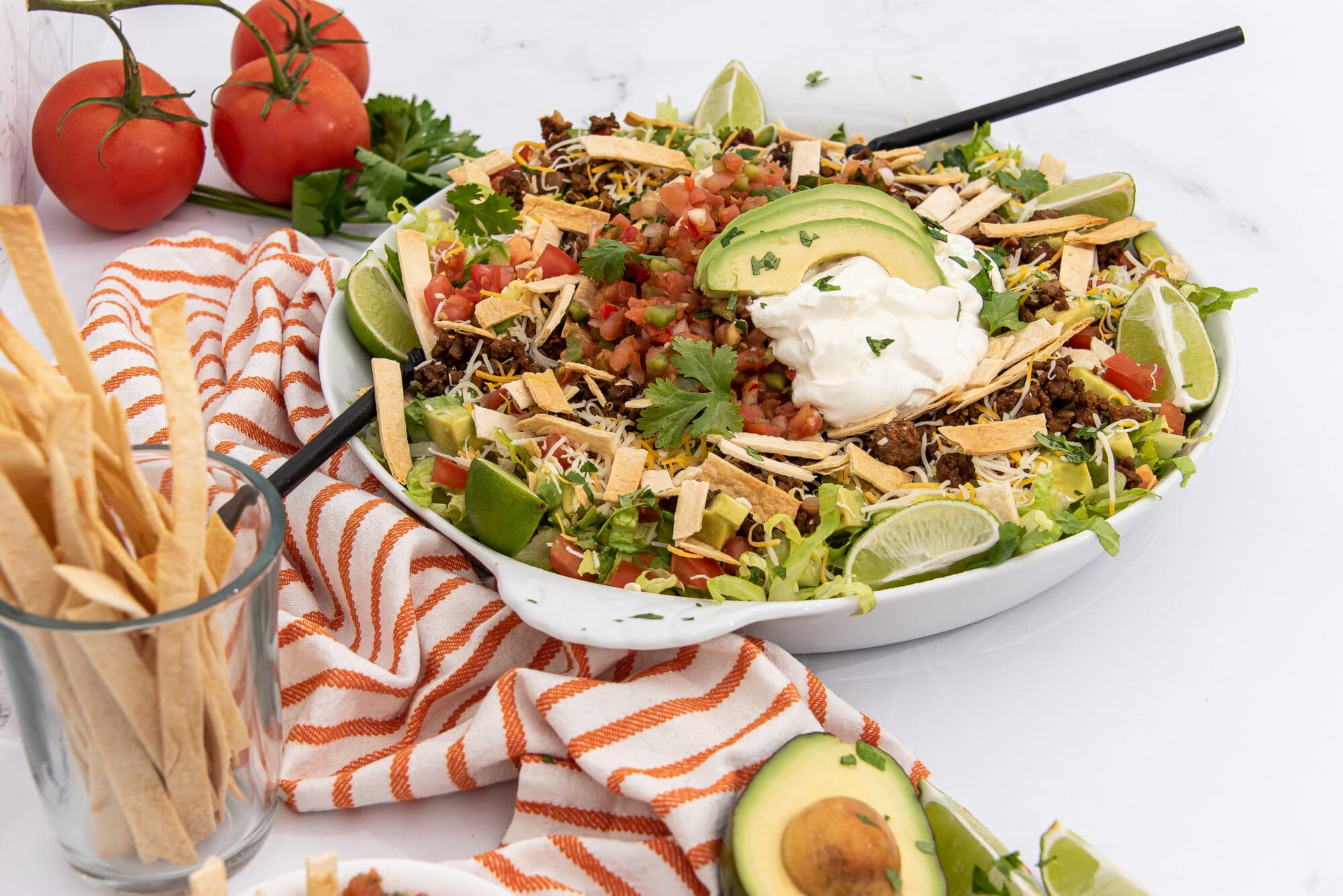 Large white bowl of taco salad containing ground beef, shredded lettuce, diced tomatoes, shredded cheese, avocado slices, and sour cream, garnished with lime wedges and cilantro. A glass of tortilla strips and fresh tomatoes are on the side, next to an orange and white striped towel, on a white countertop.