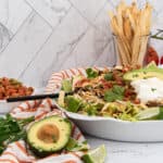 Large white serving bowl filled with taco salad, topped with ground meat, shredded lettuce, cheese, pico de gallo, sliced avocado, and sour cream. A black serving utensil rests in the bowl. A small white bowl of pico de gallo and a clear glass of tortilla strips are beside it. A halved avocado, lime wedges, and parsley are on a red and white striped towel. Fresh tomatoes on the vine and a white marble-patterned wall are in the background.