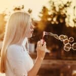 Young woman with blonde hair blowing bubbles from a red wand at sunset, with numerous iridescent bubbles floating in golden light.