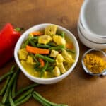 A white bowl of yellow chicken curry with green beans and orange carrots, next to a red bell pepper, scattered green beans, a small clear bowl of yellow powder, a stack of three white bowls, and two silver spoons, all on a wooden surface.
