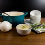 Teal cast iron pot with black ladle, white onion, white bowl of creamy soup with spoon and cilantro, three stacked white bowls, and cilantro on a checkered towel, all on a wooden surface against a dark background.