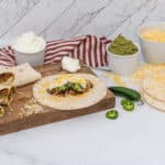 A collection of burritos and ingredients on a wooden cutting board against a white marble background. Several wrapped burritos, some cut to reveal a meat, cheese, and vegetable filling, are on the left. An open tortilla topped with ground meat, shredded cheese, sour cream, and sliced green jalapeños is also on the board. Bowls of sour cream, guacamole, and shredded cheese are visible, alongside whole and sliced jalapeños and additional tortillas. A red and white striped towel is behind the bowls.