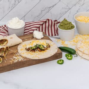 A collection of burritos and ingredients on a wooden cutting board against a white marble background. Several wrapped burritos, some cut to reveal a meat, cheese, and vegetable filling, are on the left. An open tortilla topped with ground meat, shredded cheese, sour cream, and sliced green jalapeños is also on the board. Bowls of sour cream, guacamole, and shredded cheese are visible, alongside whole and sliced jalapeños and additional tortillas. A red and white striped towel is behind the bowls.