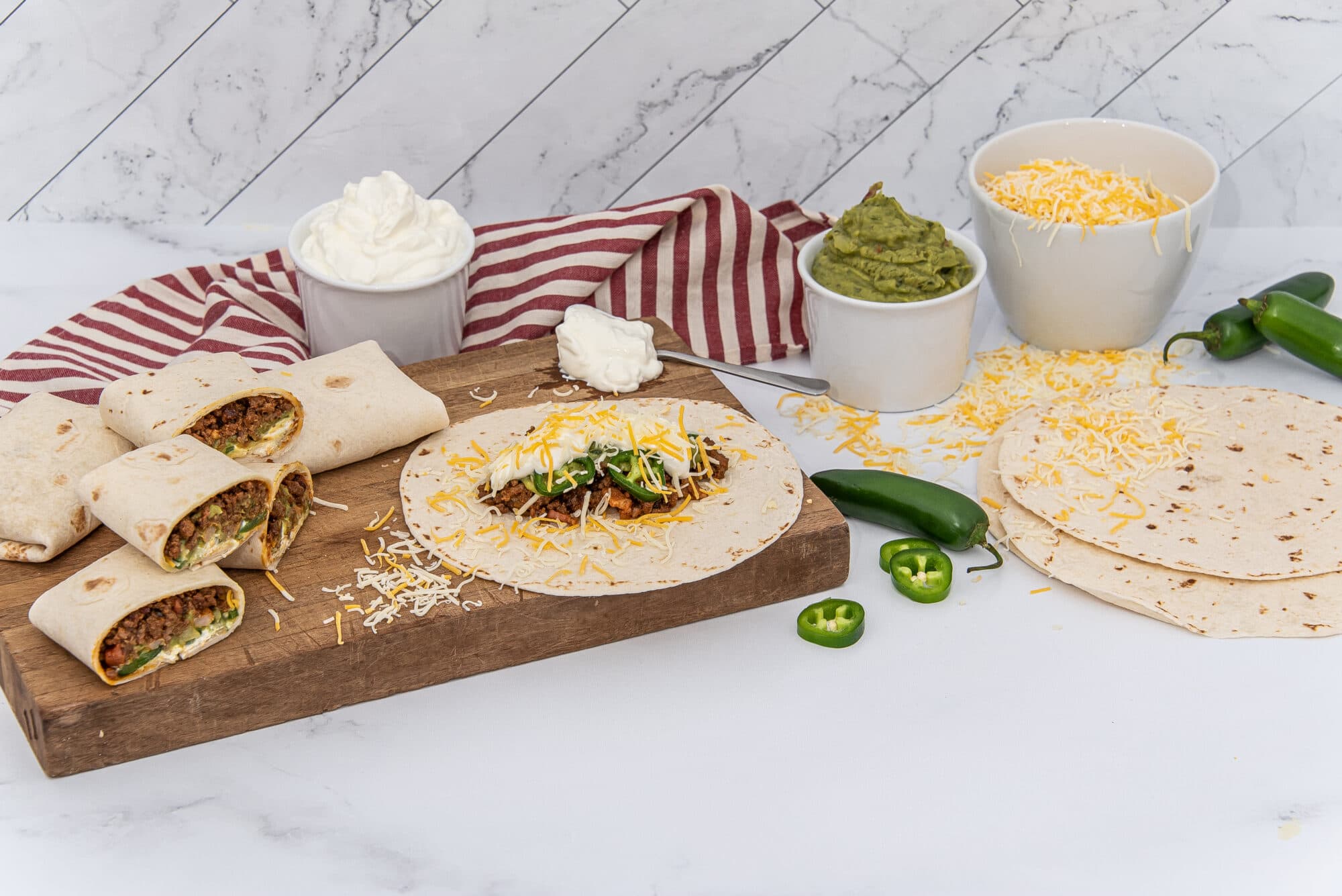 A collection of burritos and ingredients on a wooden cutting board against a white marble background. Several wrapped burritos, some cut to reveal a meat, cheese, and vegetable filling, are on the left. An open tortilla topped with ground meat, shredded cheese, sour cream, and sliced green jalapeños is also on the board. Bowls of sour cream, guacamole, and shredded cheese are visible, alongside whole and sliced jalapeños and additional tortillas. A red and white striped towel is behind the bowls.