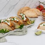 Five assembled tacos are prominently displayed on a white surface. Four tacos, on a white rectangular plate, are filled with diced chicken, avocado, and salsa, topped with white sour cream and green herbs, accompanied by lime wedges. A fifth taco, similarly prepared, rests on a white napkin. In the background, whole avocados, a stack of plain tortillas, red tomatoes on a vine, and a clear bowl of diced chicken are visible. The setting includes a white counter with subtle grey veining and a green and white striped towel.