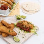 Fried fish fillets and fish tacos with lettuce, salsa, and lime wedges, presented on white serving dishes and a cutting board against a white surface.