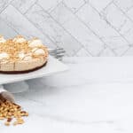 Round light brown cake with a dark crust, topped with white cream swirls, caramel drizzle, and peanuts. It sits on a white cake stand. A white paper bag spills peanuts onto a white marble counter, with stacked plates and silver forks nearby. A white marble-patterned wall is in the background.