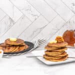 Two plates of golden brown pancakes with amber syrup. One plate holds a tall stack topped with a pat of yellow butter and orange pumpkin mash. The other has a smaller stack with butter. A clear glass bottle of syrup, a white bowl of orange pumpkin mash, and a white bowl of yellow butter are arranged on a white marble surface. A navy and white striped napkin is between the plates. The background is a white marble herringbone tiled wall.