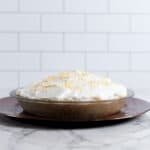 A pie with a brown filling in a clear glass dish, topped with white whipped cream and sprinkled with toasted coconut flakes. It sits on a round reddish-brown metallic plate over a white marble surface, with a white subway tile wall in the background.