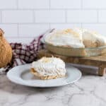Coconut cream pie in a glass dish with one slice on a white plate, topped with whipped cream and toasted flakes. A whole coconut and red plaid cloth are alongside. The scene is set on a wooden board, white marble counter, and white tiled wall.