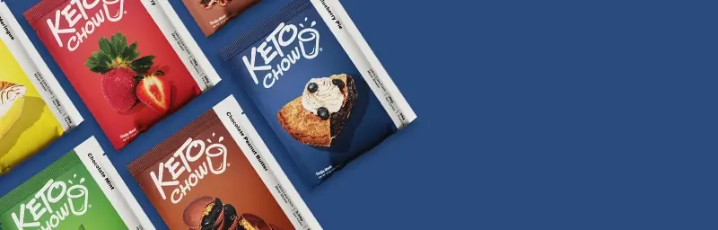 A variety of Keto Chow single-serving meal mix packets arranged diagonally against a royal blue background. Visible flavors include Lemon Meringue, Strawberry, Chocolate Mint, Chocolate Peanut Butter, and Blueberry Pie.