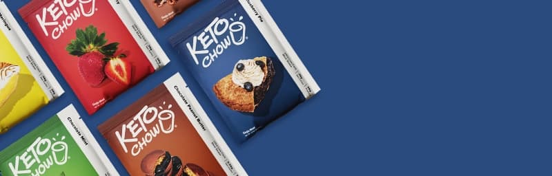 A variety of Keto Chow single-serving meal mix packets arranged diagonally against a royal blue background. Visible flavors include Lemon Meringue, Strawberry, Chocolate Mint, Chocolate Peanut Butter, and Blueberry Pie.