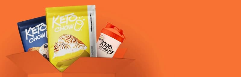 Keto Chow subscription bundle with two colorful meal mix bags—one in bright yellow labeled Lemon Meringue and a second navy blue bag with an unreadable flavor—alongside a white Keto Chow shaker bottle with an orange lid, all emerging from an orange box against a bold orange background.