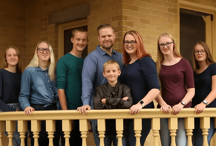 A family of eight, including six children and two adults, stands on a porch with a beige railing.