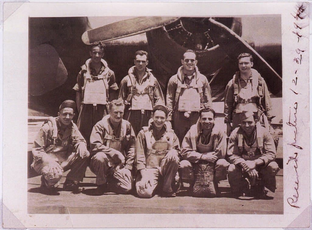 Sepia-toned group portrait of ten men in flight suits and harnesses, with an airplane propeller and engine in the background. Four men stand in the back row, and six men crouch in the front row. Handwritten text is visible vertically on the right edge of the photograph.