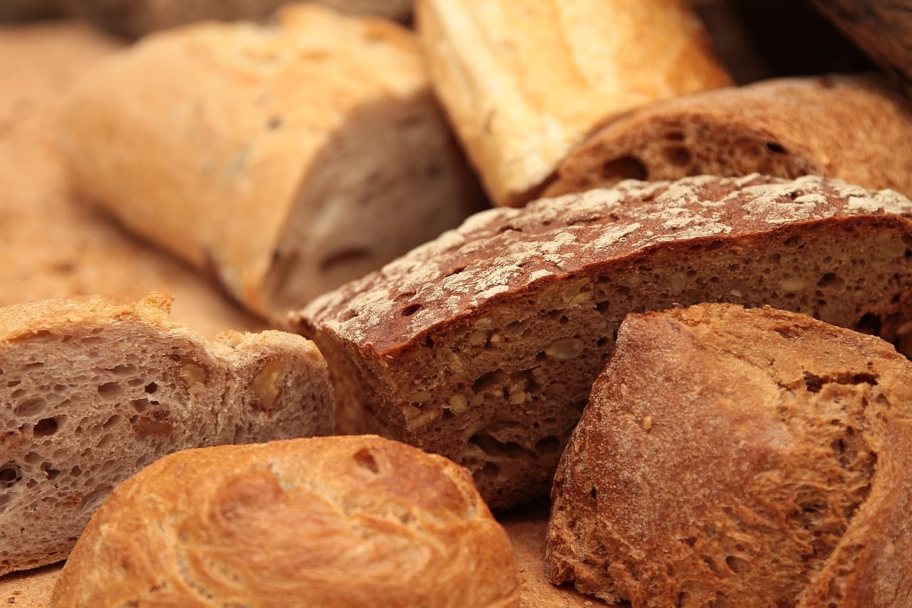 Various artisan breads, including sliced and whole loaves, in shades of golden brown to dark reddish-brown. Visible bread interiors show open crumb textures with nuts or seeds, and some crusts are dusted with flour.